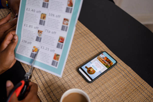 An unrecognizable woman cutting out food coupons with scissors while sitting at a dining table at home in Newcastle Upon Tyne, England. She is trying to save money during the cost of living crisis. Her phone is unlocked and showing an internet page for a food subscription service.