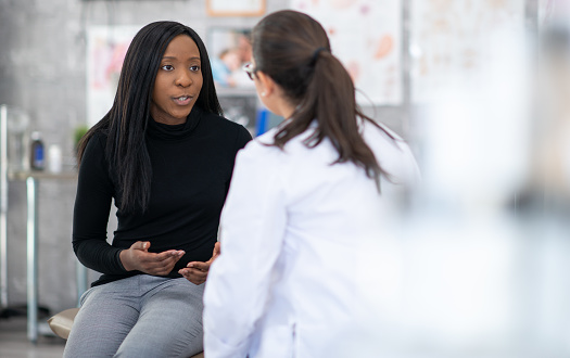 A middle aged woman of African decent, sits up on an exam table as she talks with her doctor about her health concerns. She is dressed casually and her female doctor is seated across from her in a white lab coat as the two carry on a discussion.