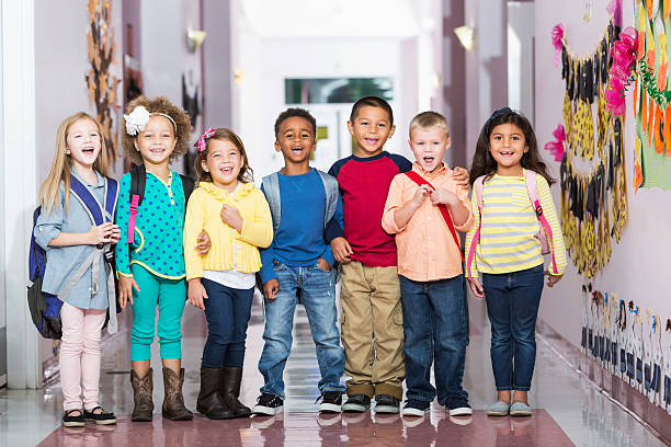 A multi-ethnic group of seven children standing in a row in a school hallway, laughing and smiling at the camera. The little boys and girls are kindergarten or preschool age, 4 to 6 years.